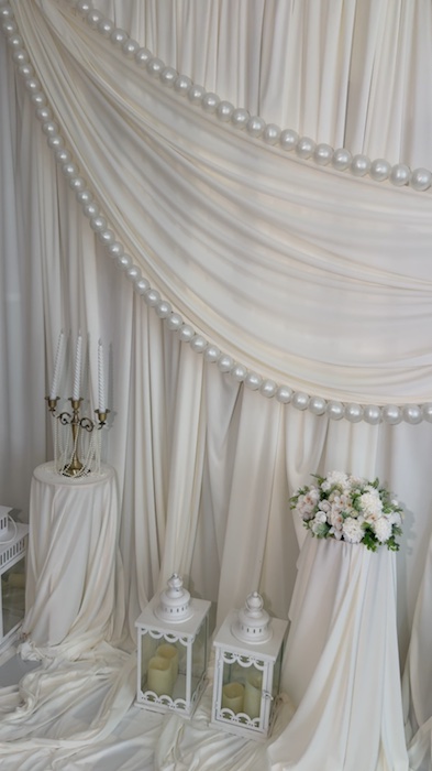 Ivory draped backdrop edged with pearl-like strands, a floral arrangement, white lanterns and a candelabra.