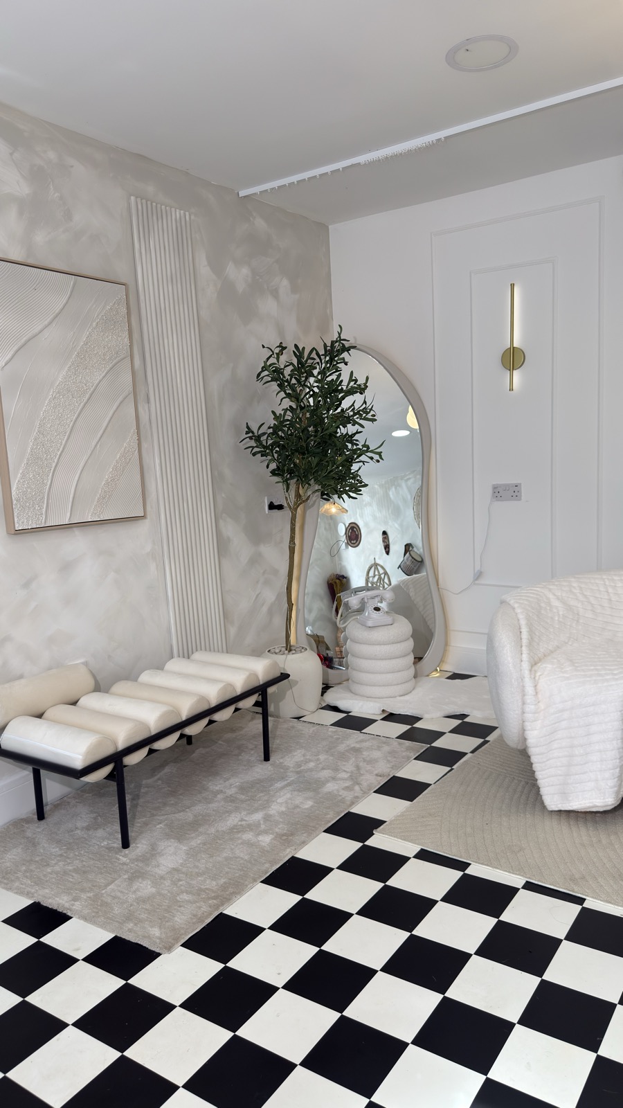 Arched floor mirror beside a leafy tree and white vintage-style telephone, with cream bench, wall art and checkerboard flooring.
